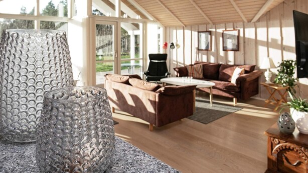 Een ruime woonkamer met een houten plafond, een bruine zithoek, een witte salontafel en een groot raam met uitzicht op de omgeving.
