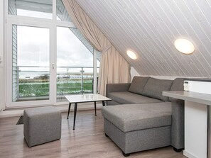 A modern interior with a grey sofa, ottoman, and table, a large window with a view of a body of water, and a sloped ceiling with built-in lighting.