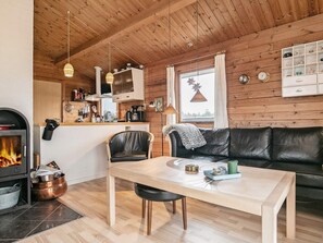 A cozy living room with a fireplace, a wooden ceiling, a black leather sofa, a wooden dining table, and a kitchen area with a window. - 6 Person Holiday Home in Hirtshals (Hirtshals)