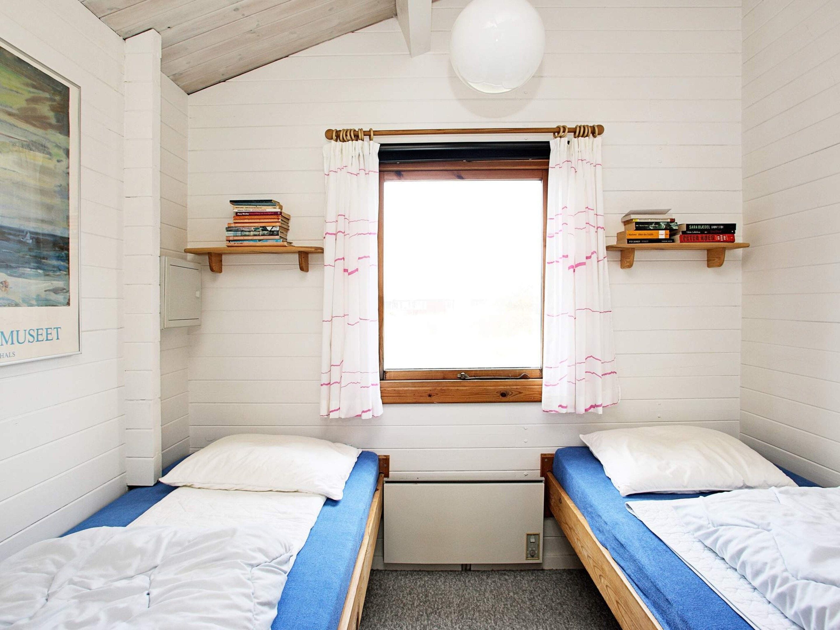 A small, cozy room with two single beds, a window with curtains, and wooden shelves with books.