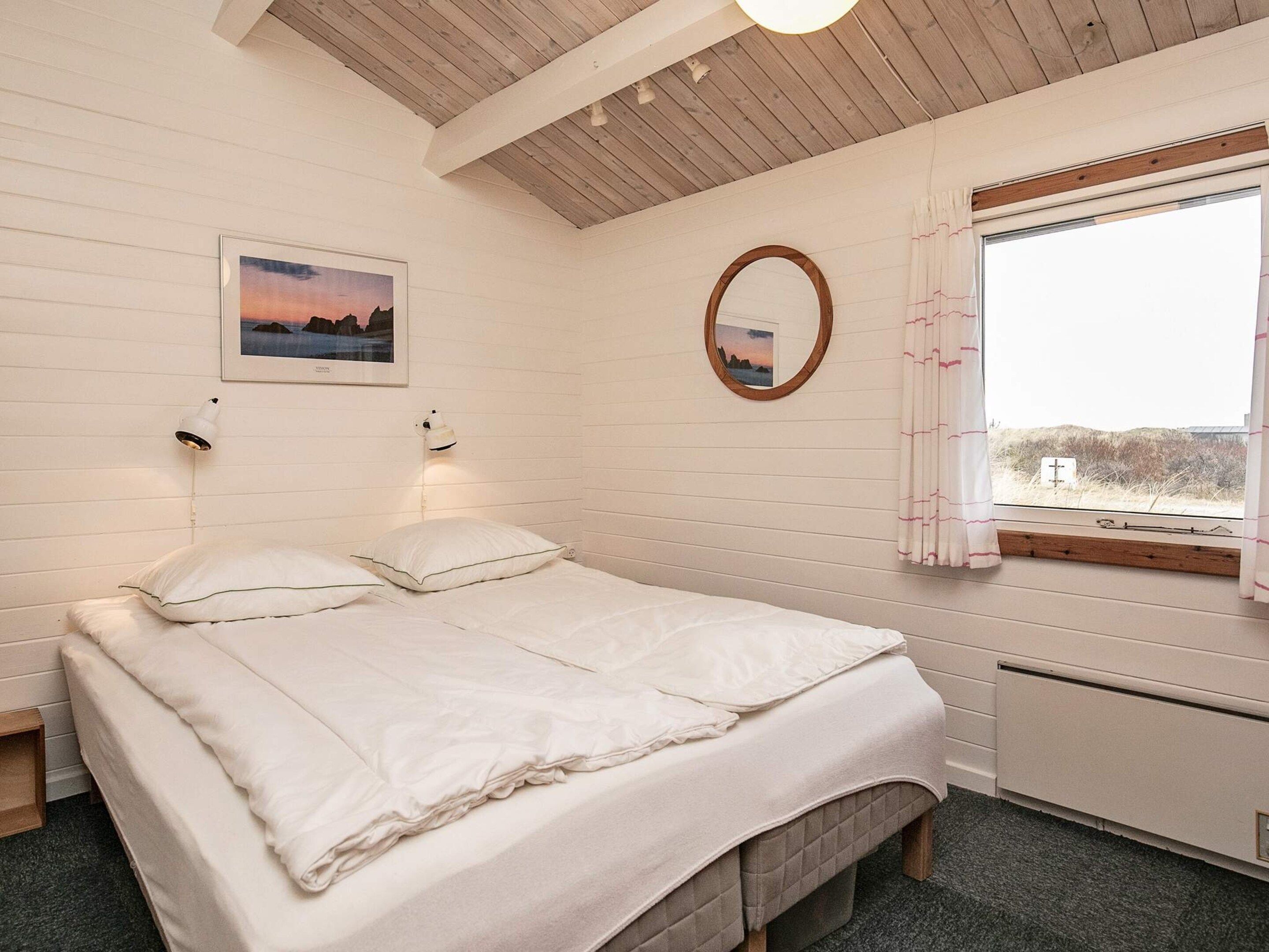 A bedroom with a bed, a bedside table, a window with curtains, a wall-mounted mirror, and a framed picture.