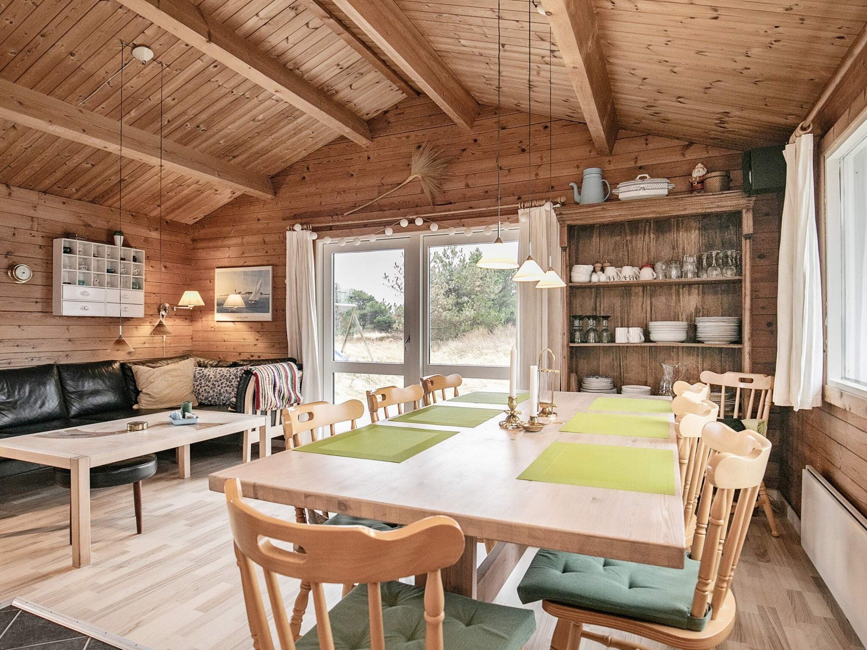 A cozy cabin interior with a wooden dining table, chairs, a sofa, and a kitchen area with shelves.