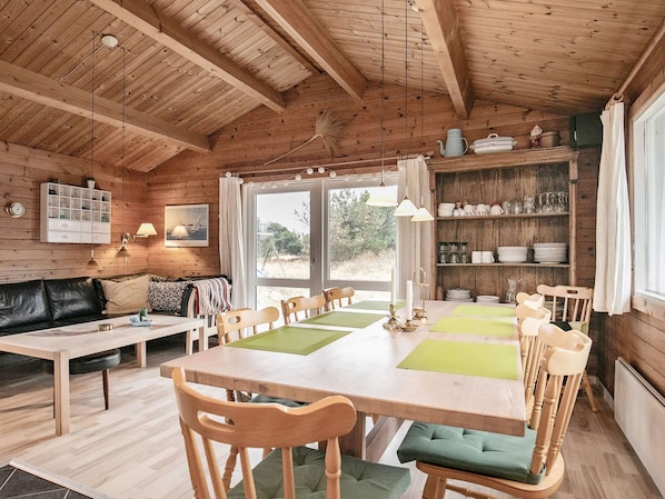 A cozy cabin-style room with a wooden ceiling, a dining table with chairs, a sofa, and a bookshelf.