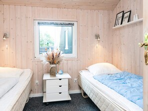 A room with two beds, a window, a bedside table with a vase, and a wall-mounted shelf.