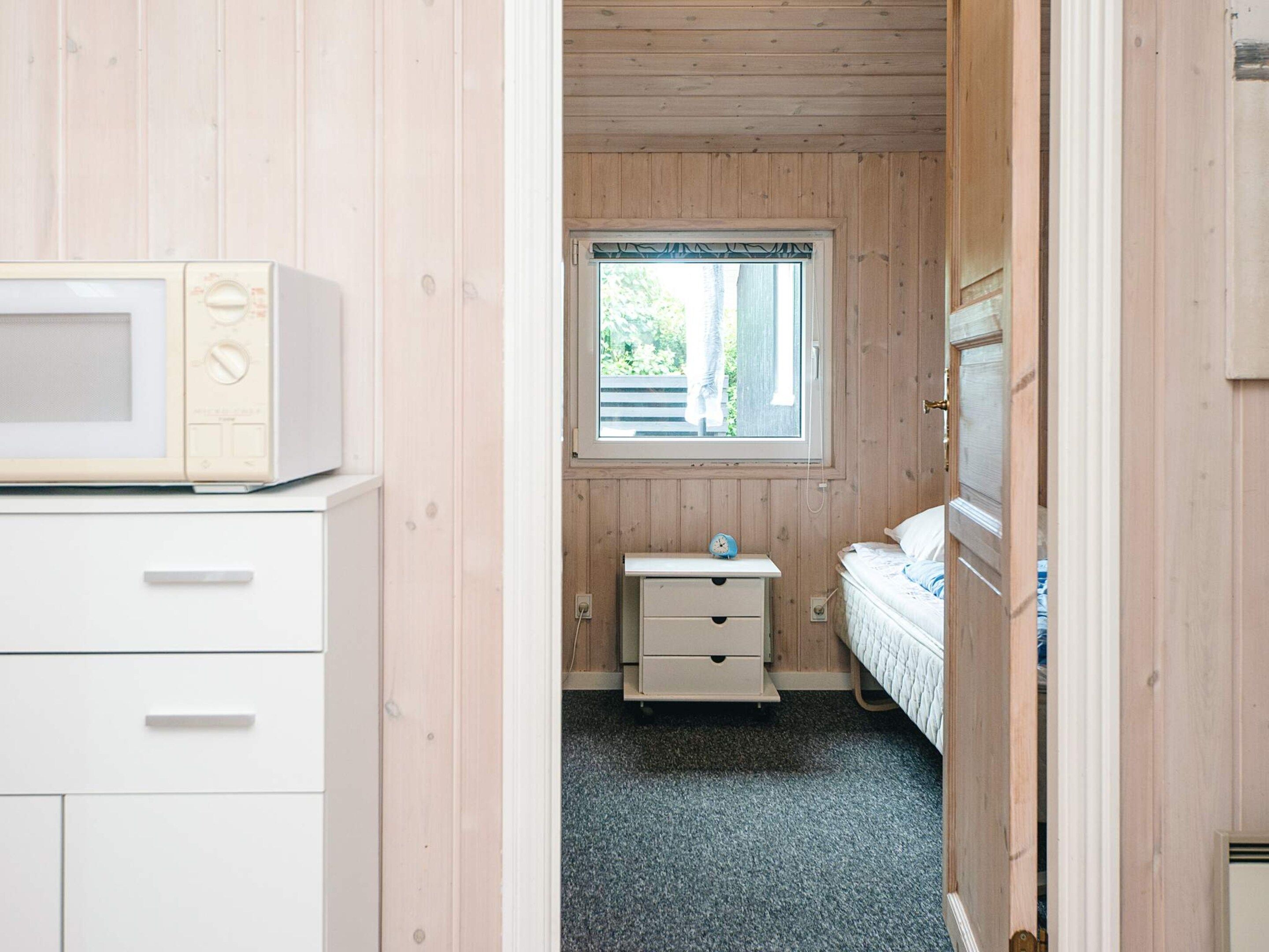 A bedroom with a wooden floor, a window with a view of greenery, a bed, a bedside table with a clock, and a microwave on a cabinet.