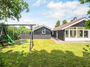 Un jardin avec une balançoire, un toboggan, une maison avec un toit rouge et une zone délimitée par une clôture.