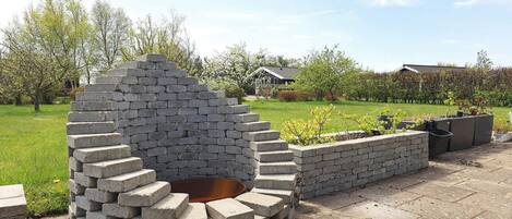 A circular outdoor fire pit made of stacked bricks, surrounded by a low brick wall, set in a grassy area with trees and a house in the background.