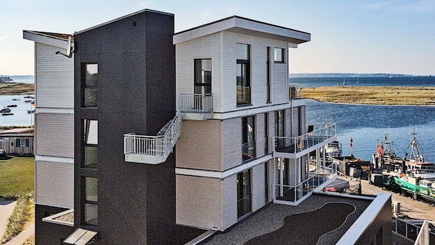 Un immeuble moderne à plusieurs étages, doté d’un balcon, de grandes fenêtres et offrant une vue sur l’eau et les bateaux.