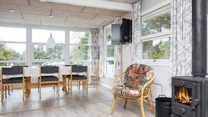 Une pièce chaleureuse avec une table à manger, des chaises, un fauteuil en osier, une cheminée et de grandes fenêtres.