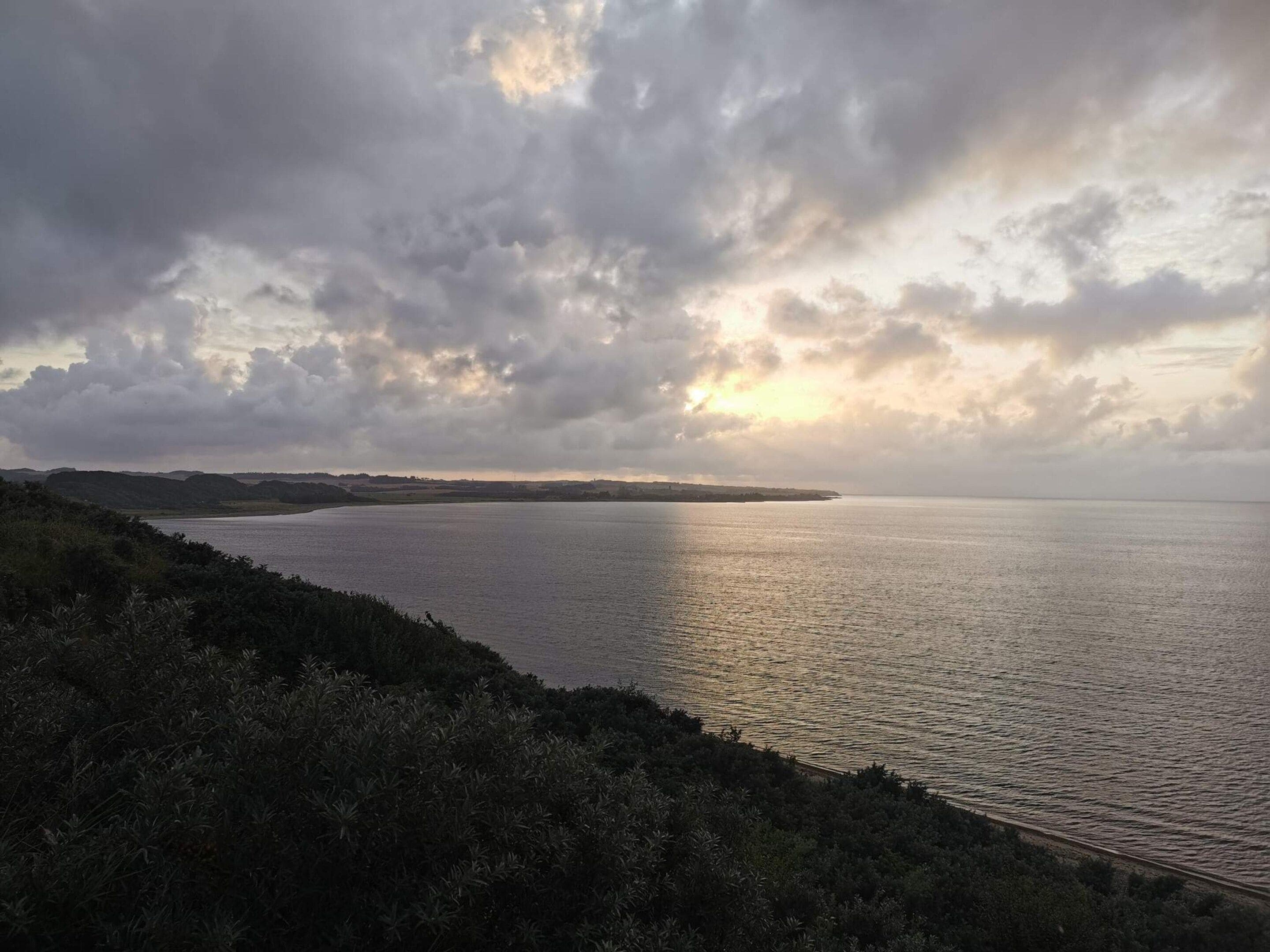 A coastal landscape with a body of water, vegetation, and a cloudy sky.