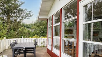 Una terraza de madera con mesa y sillas, cerca viva blanca y grandes ventanales.
