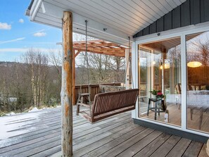 Une terrasse en bois avec une balançoire, une porte vitrée donnant sur l’intérieur et une vue sur les arbres et les collines.