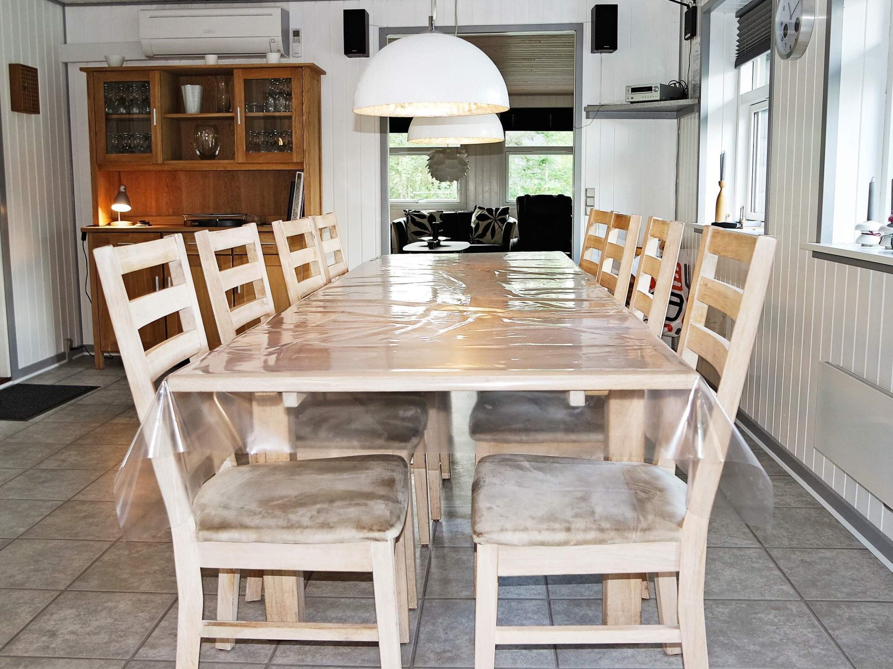 Een ruime eethoek met een lange tafel en houten stoelen, een keuken op de achtergrond, en een groot raam dat natuurlijk licht binnenlaat.
