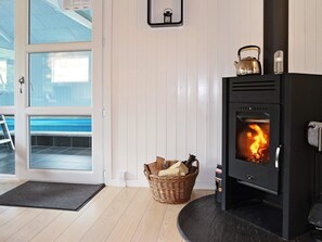 Una habitación con estufa de leña, una cesta de mimbre con leña y vista a una piscina a través de un ventanal.