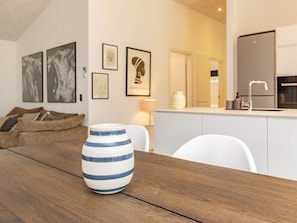 Ein modernes Wohnzimmer mit Holztisch, weißem Sofa und einer Küchenzeile im Hintergrund.