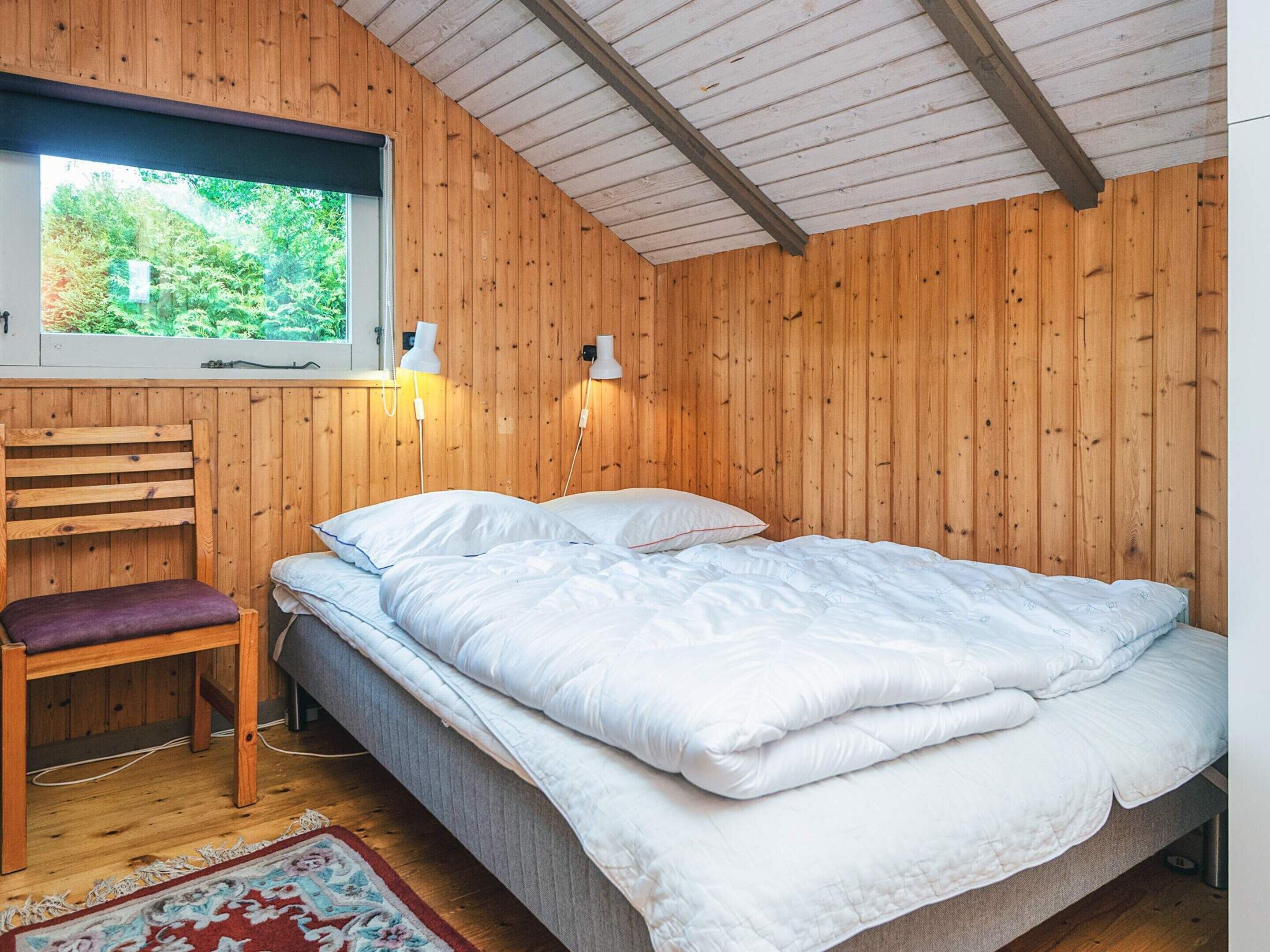 A bedroom with a bed, a chair, a window, and a wooden ceiling.