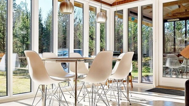 A modern dining area with a large table and white chairs, large windows, and a wooden ceiling. - 6 Person Holiday Home in Norre Nebel (Norre Nebel)