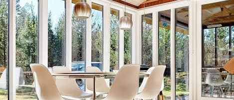 A modern dining area with a large table and chairs, large windows, and a wooden ceiling structure.