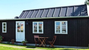 A black wooden house with a white door and windows, a small patio with two wooden chairs, and a lawn. - 4 Person Holiday Home in Skagen (Skagen)