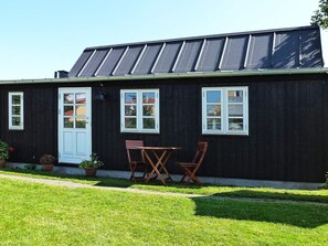 Une maison en bois peinte en noir, avec une porte et des fenêtres blanches, un petit jardin avec de la pelouse et des plantes en pot, ainsi qu’une table et des chaises en bois à l’extérieur.