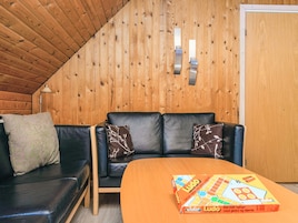 Una habitación acogedora con techo de madera, un sofá de cuero negro con cojines estampados, una mesa de madera y un juego de Ludo.
