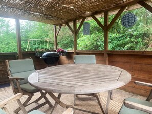 Een houten terras met een ronde tafel en stoelen, omringd door groen.