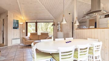 Ein gemütliches Blockhaus-Interieur mit einem runden Esstisch, weißen Stühlen, einer Couch und einer Küchenzeile.