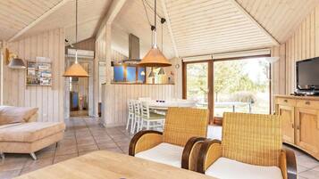 A spacious living area with a high ceiling and wooden walls, a dining area with wicker furniture, and a kitchen with wooden cabinets.