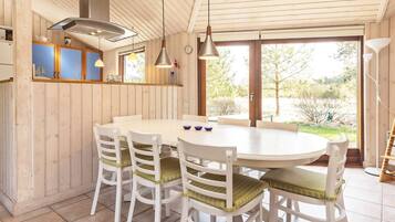 A wooden interior with a round dining table, white chairs with green cushions, pendant lights, and a view through large glass doors.