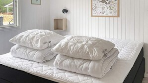 Ein ordentlich bezogenes Bett mit einer gesteppten weißen Bettdecke und Kissen, ein Nachttisch mit einer Lampe und ein gerahmtes Pflanzenbild an der Wand.