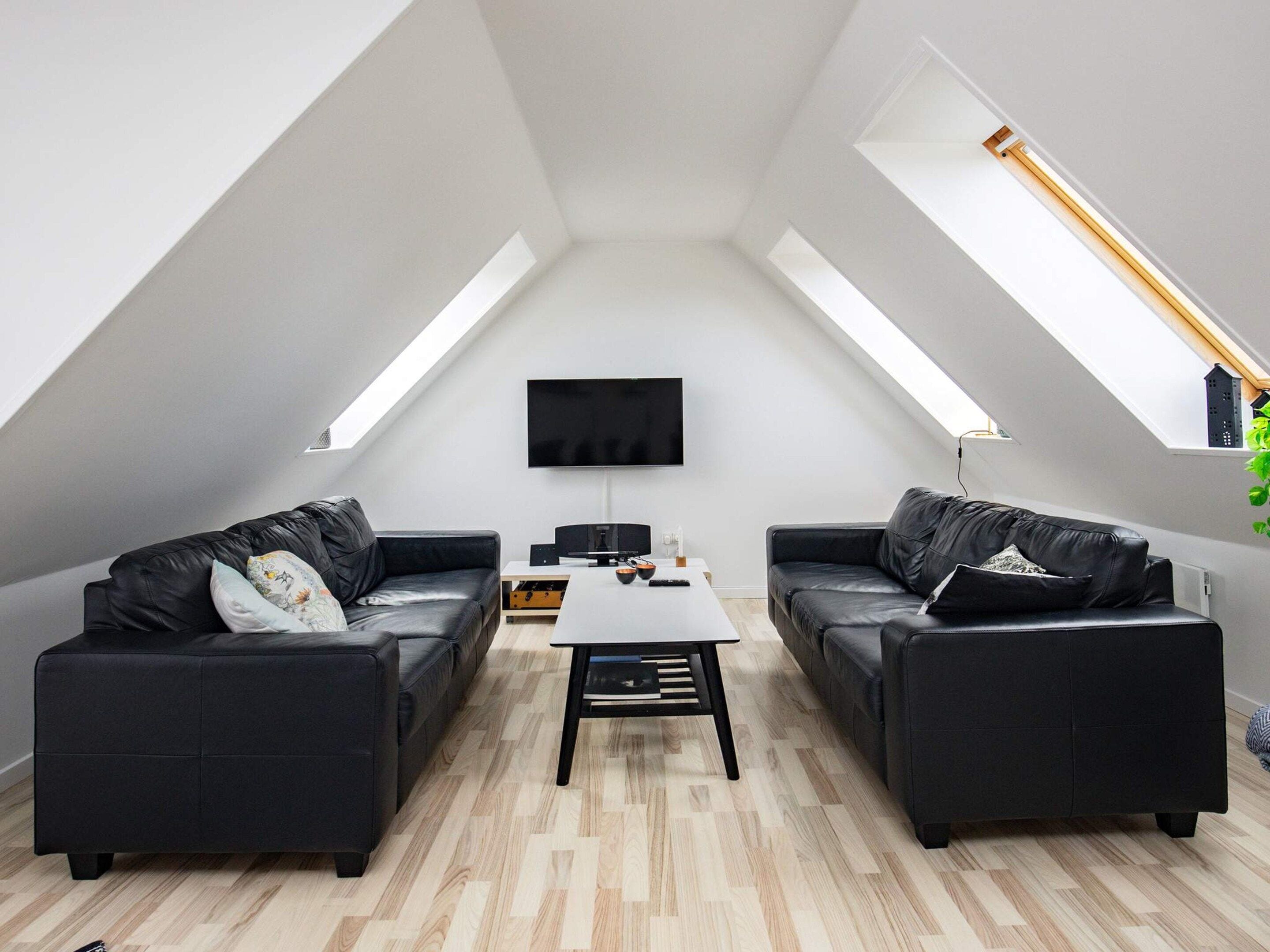 Ein modernes Schlafzimmer im Loftsstil mit schrägem Dach, zwei schwarzen Ledersofas, einem zentralen Couchtisch und einem an der Wand montierten Flachbildfernseher.