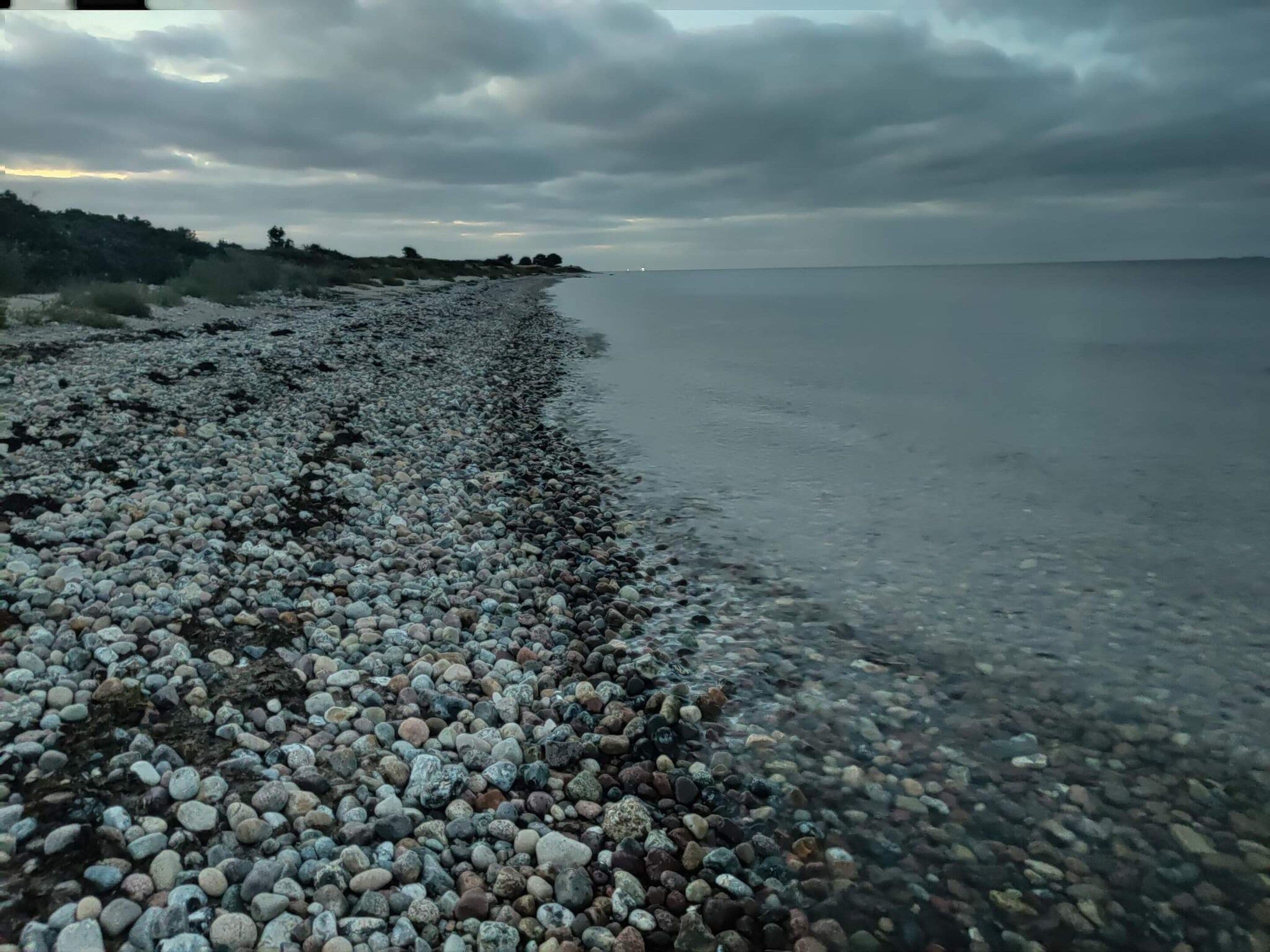 A pebbled beach with a calm sea and overcast sky.