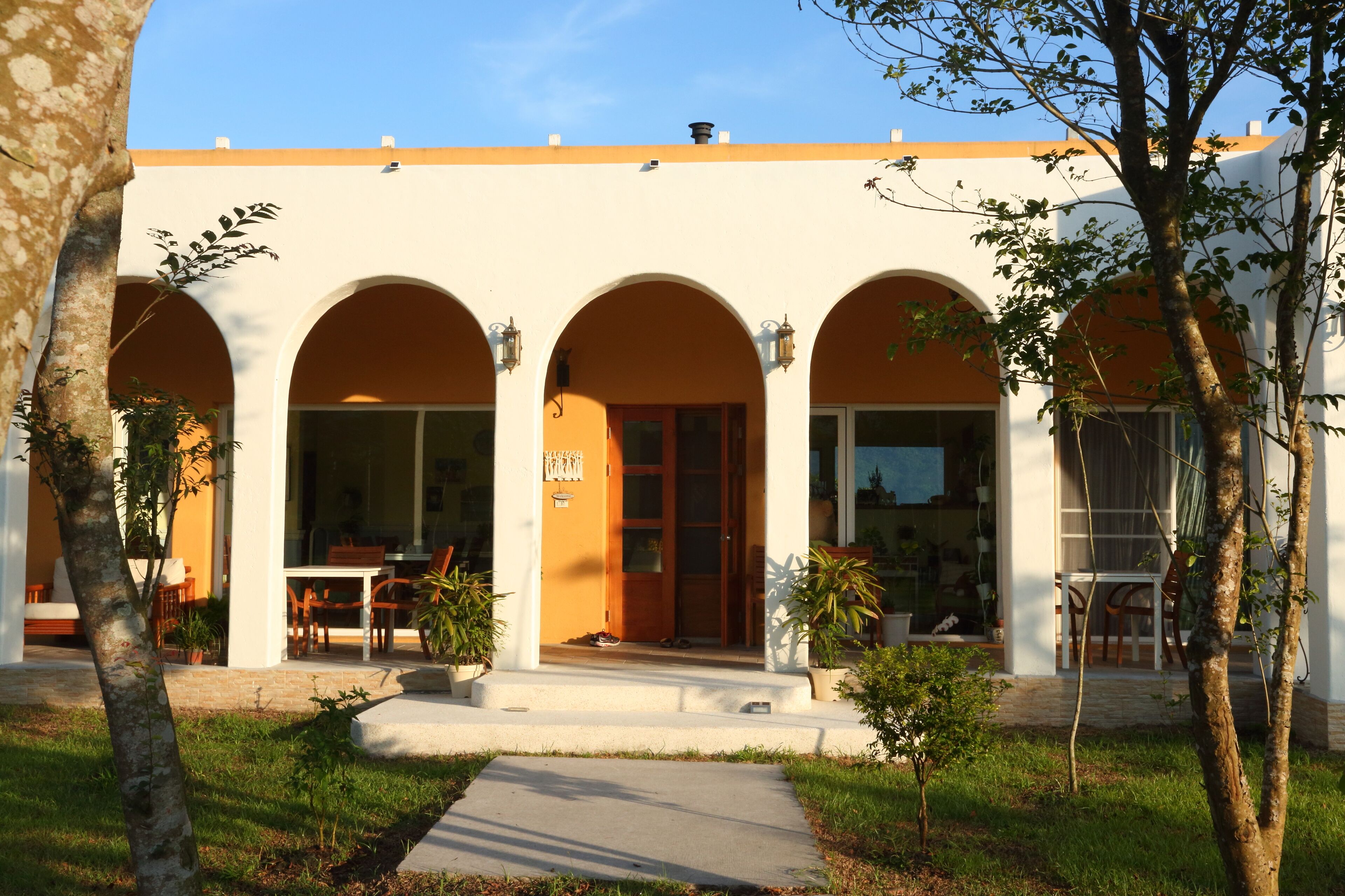 A building with three arched doorways, a covered patio area, and a small garden with trees and potted plants.