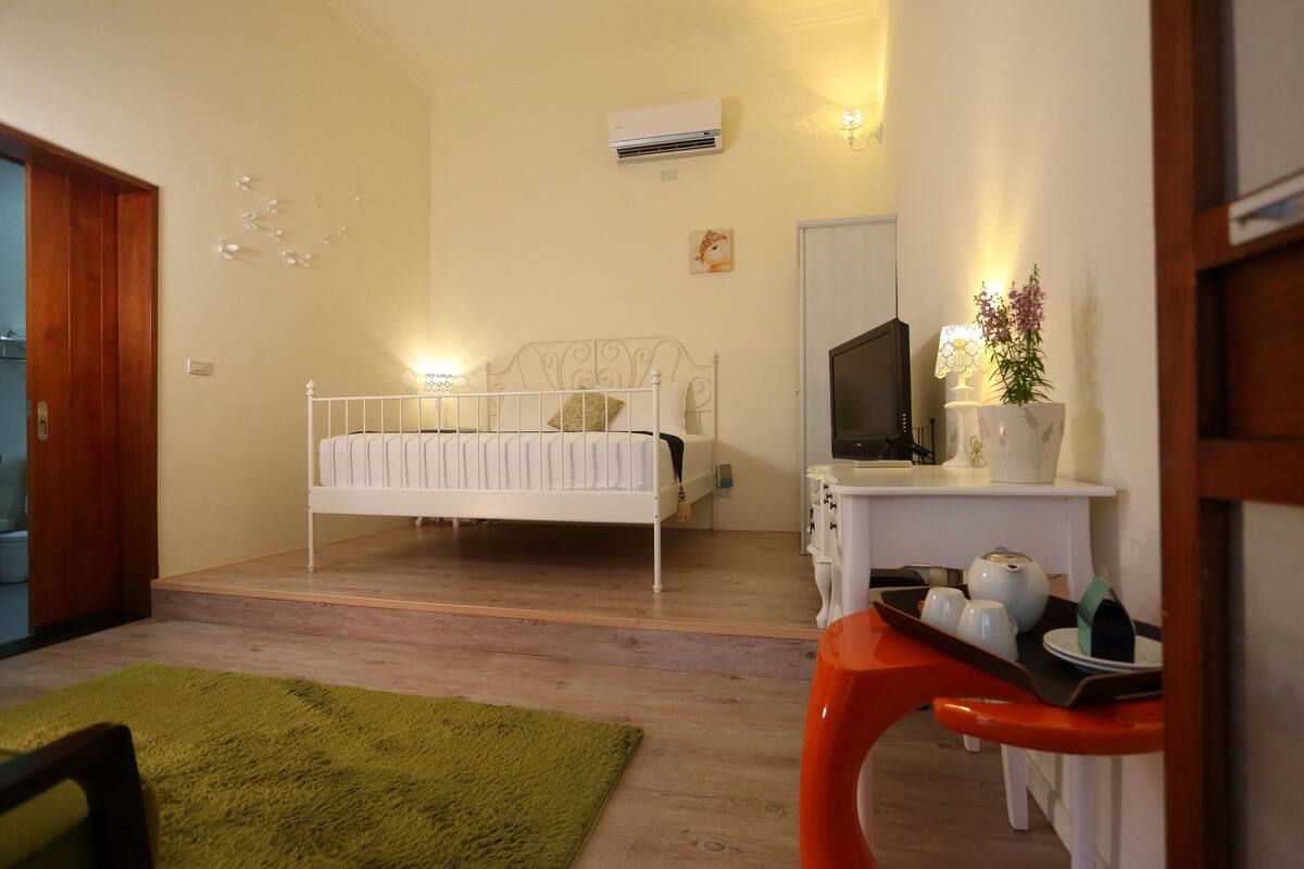 A bedroom with a bed, a desk with a television, a small table with a vase, and a green rug.