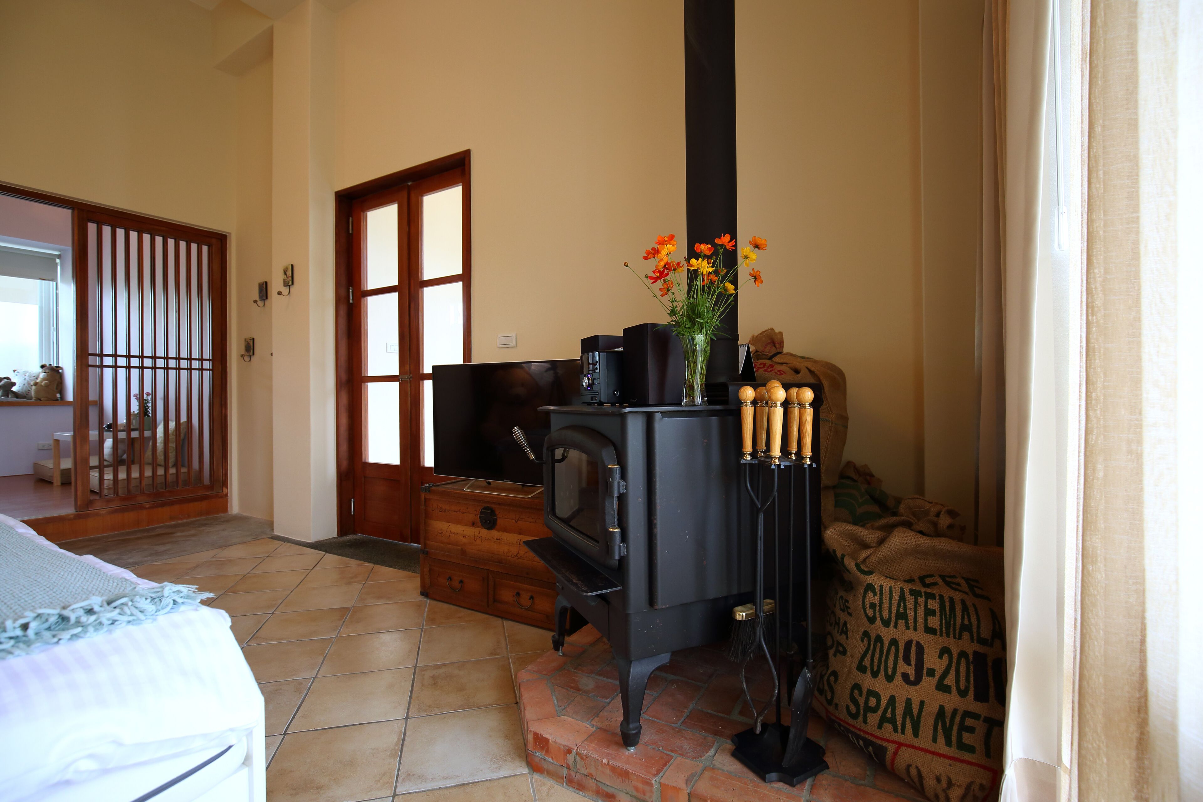 A room with a bed, a wooden chest, a black stove, and a vase with flowers.