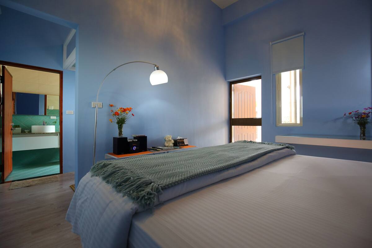 A bedroom with a large bed, a bedside table with a lamp, a window, and a view of a bathroom.