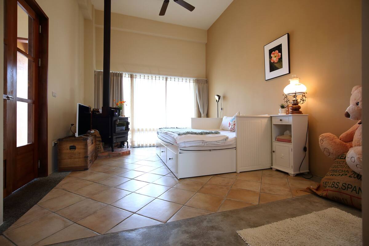 A bedroom with a bed, a bedside table with a lamp, a dresser, a large stuffed animal, and a window with curtains.