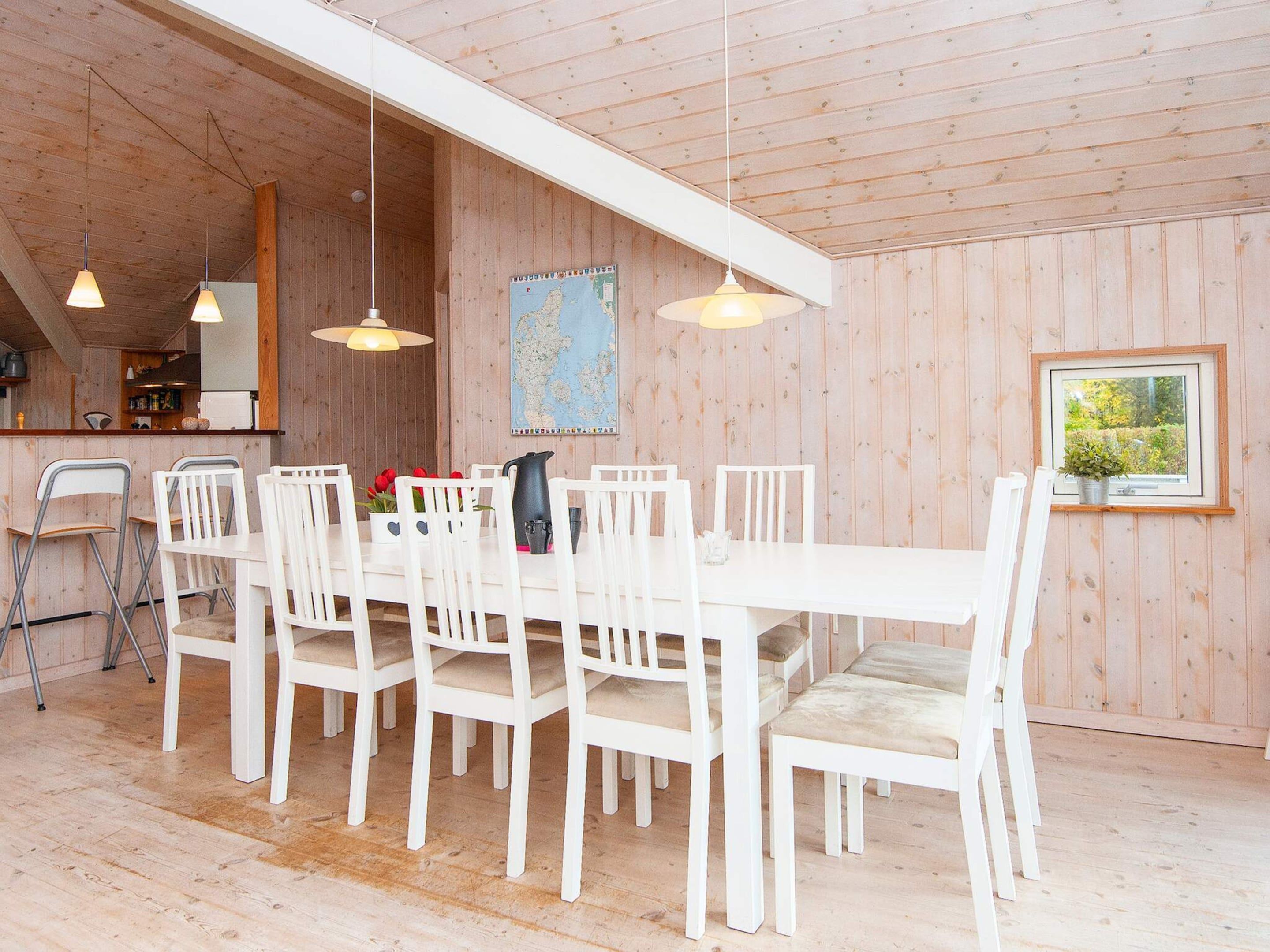A dining area with a long table and white chairs, wooden walls, and hanging pendant lights.