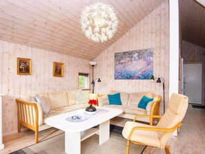 Amplia habitación con techo de madera, un sofá blanco, una mesa de centro y una silla. Hay un cuadro en la pared y una ventana con vistas.