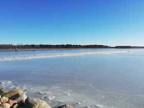 Ein zugefrorener See mit klarem Eis, umgeben von Bäumen und einem Gebäude in der Ferne.