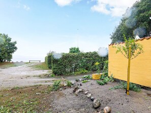 Un sendero de tierra que lleva a un banco, una pared amarilla y un seto con humo elevándose.