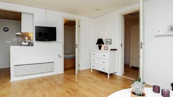 Quarto moderno com televisão de tela plana na parede, penteadeira branca com gavetas, pequena mesa com planta em vaso e piso de madeira.