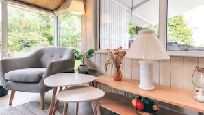 Una habitación acogedora con un sillón gris, una mesita redonda, una lámpara blanca y una estantería de madera con un jarrón y un frutero.