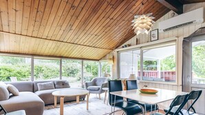 Una habitación amplia con techo de madera, un ventanal grande, un sofá, una mesa de comedor con sillas y una lámpara colgante.