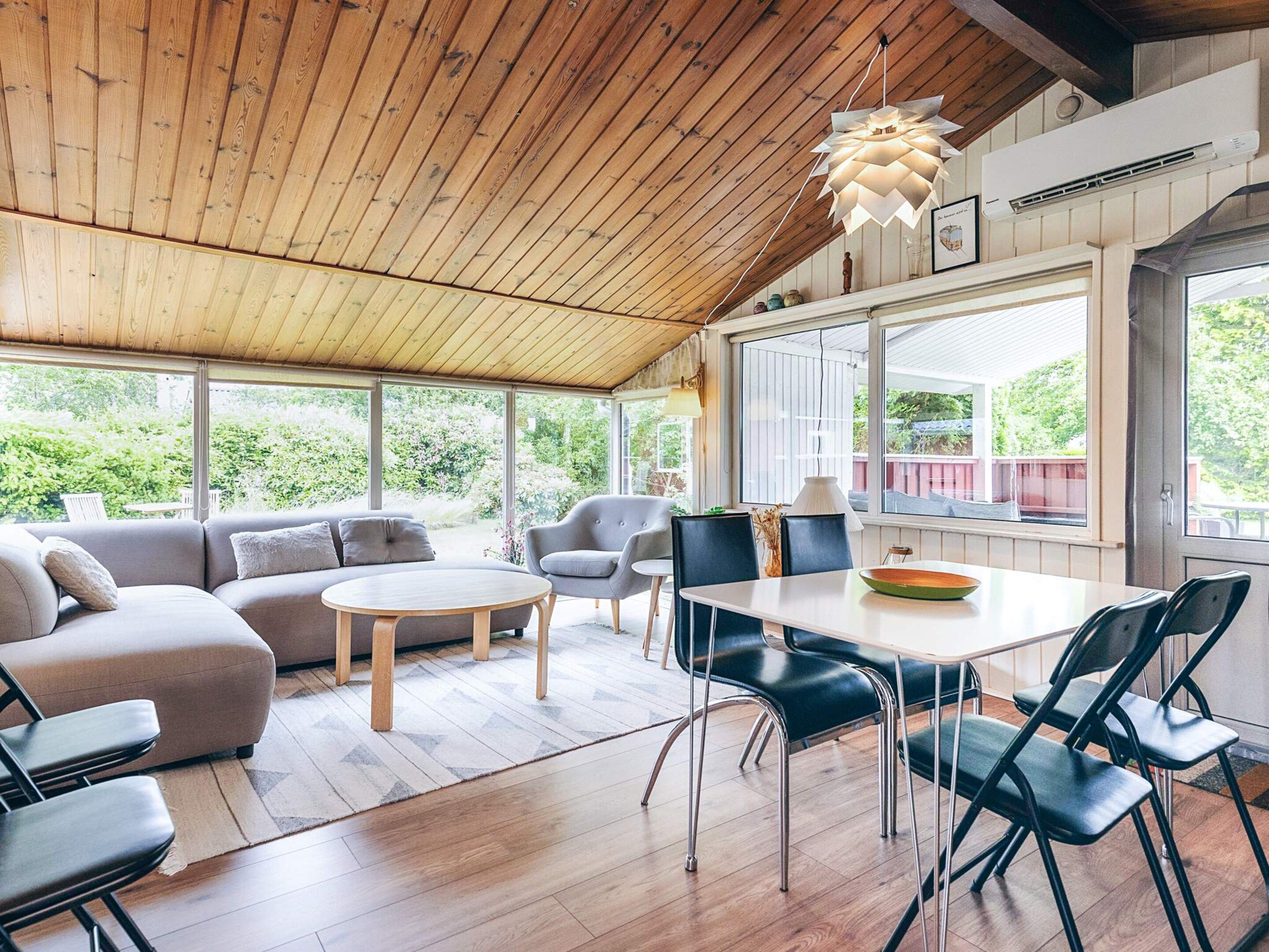 Een ruime kamer met een houten plafond, een eettafel met stoelen, een bank en een groot raam met uitzicht op groen.