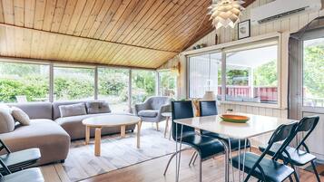 Een ruime kamer met een houten plafond, een eettafel met stoelen, een bank en een groot raam met uitzicht op groen.