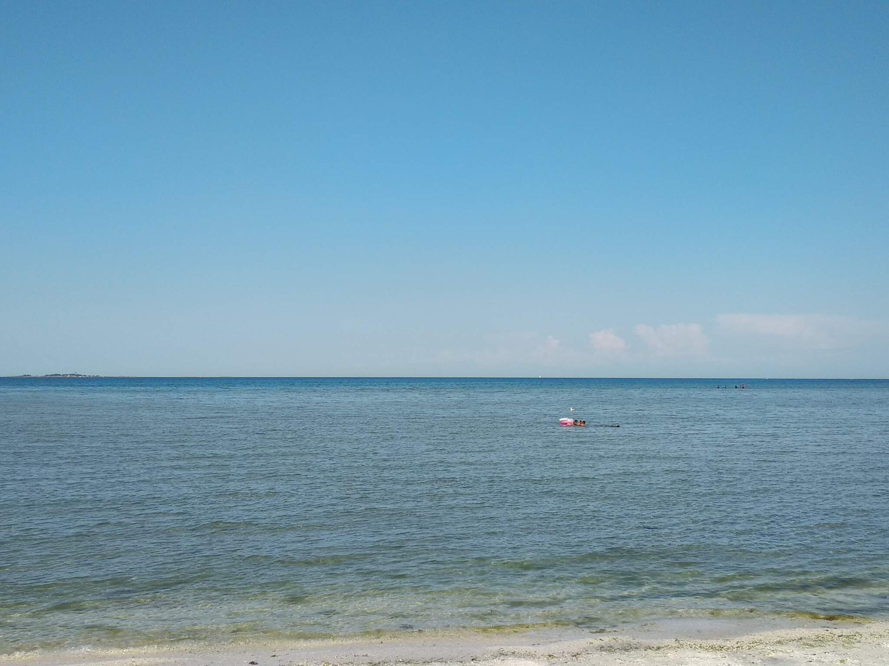 Een strand met helder water, een eiland in de verte en iemand die zwemt.