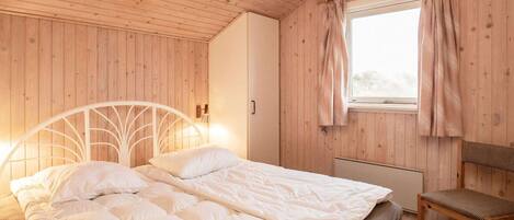 Un dormitorio acogedor con techo y paredes de madera, una cama con ropa blanca, una mesita de noche con lámpara y una ventana con cortinas.