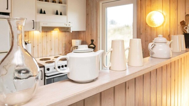 A kitchen with wooden cabinets, a stove, white mugs, and a clear glass pitcher. - 4 Person Holiday Home in Blokhus (Blokhus)