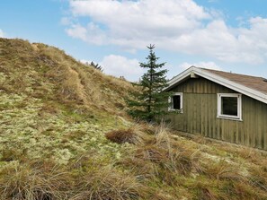 A small wooden cabin with a single window, surrounded by grassy terrain and a coniferous tree. - 4 Person Holiday Home in Blokhus (Blokhus)